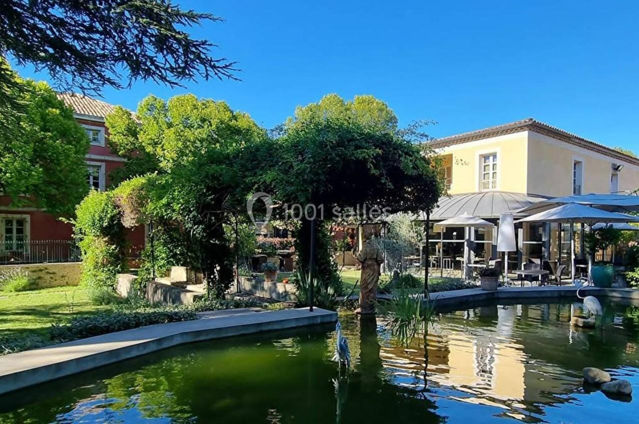 Jardin avec bassin, végétation luxuriante, terrasse ombragée et bâtiments en arrière-plan sous un ciel bleu.