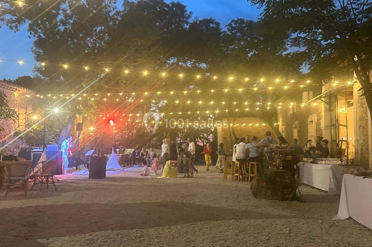 Cour extérieure éclairée par des guirlandes lumineuses, avec des tables, des convives et une scène en arrière-plan.