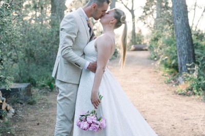 Un couple en tenue de mariage marche main dans la main devant un bâtiment en pierre et des arbres.