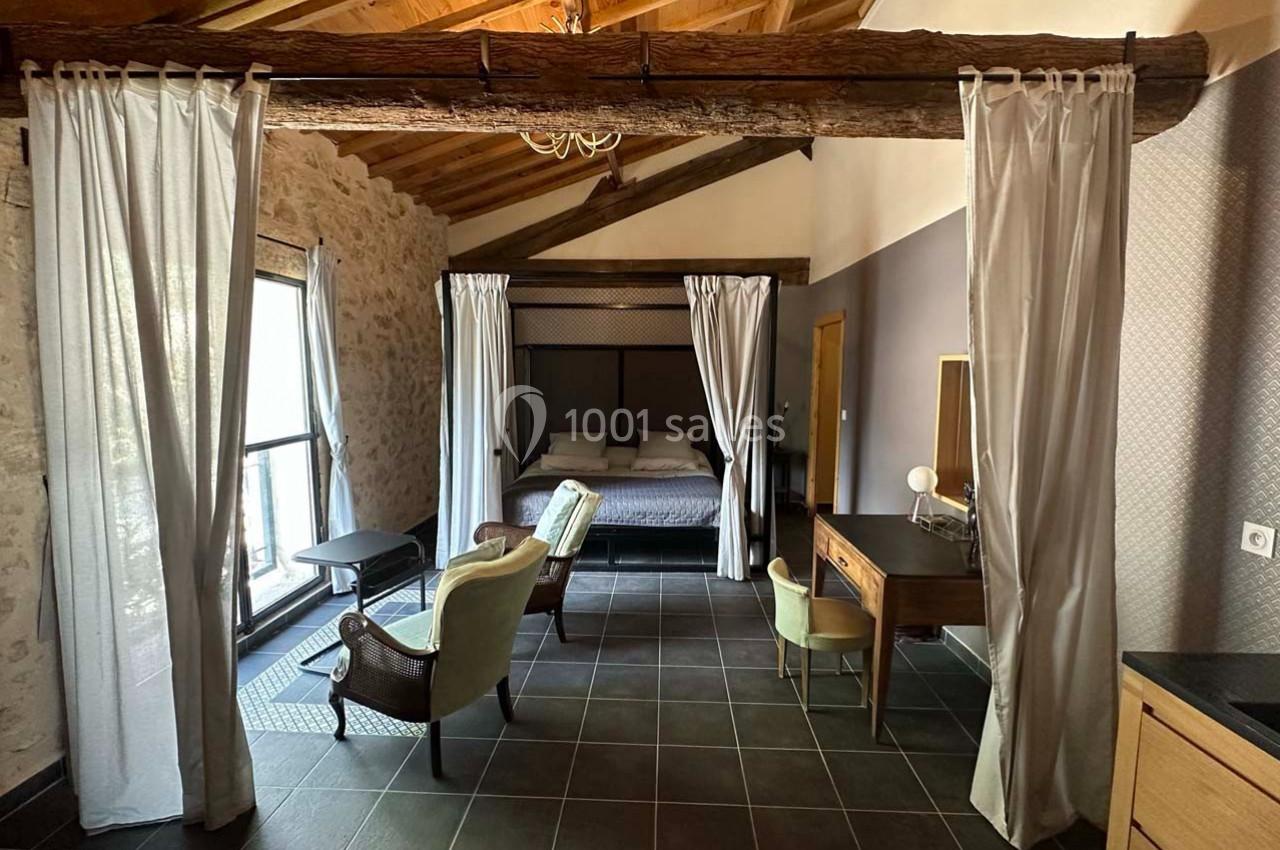 Chambre avec lit à baldaquin, murs en pierre, plafond en bois, mobilier ancien et grande fenêtre laissant entrer la lumière.