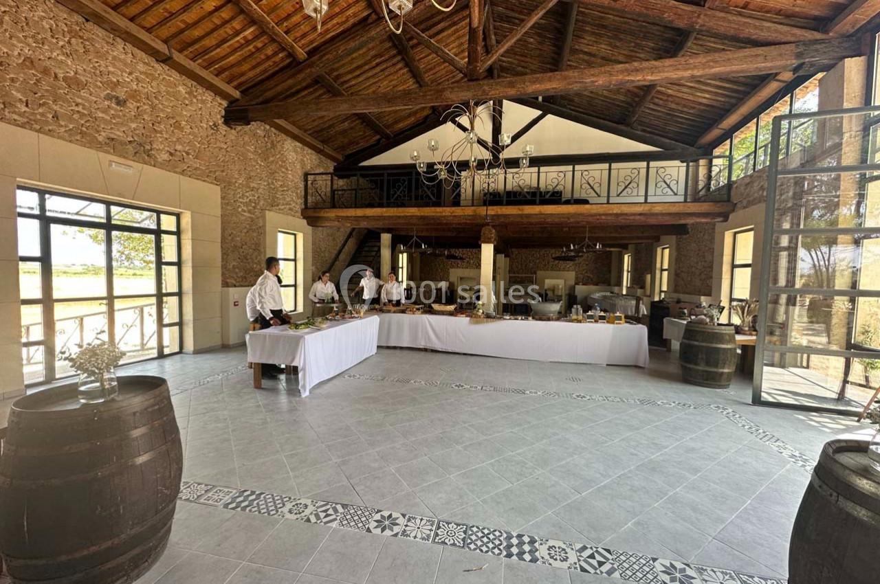 Salle spacieuse avec murs en pierre, plafond en bois, grandes fenêtres et tables dressées pour un buffet.