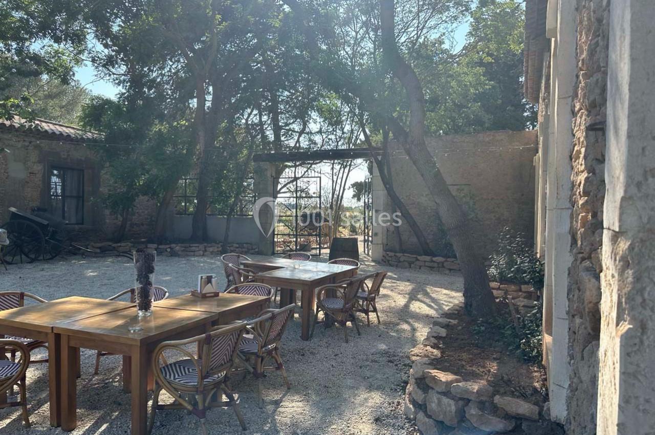 Cour extérieure ensoleillée avec tables en bois, chaises en rotin, arbres et vue sur un portail ouvert en pierre.