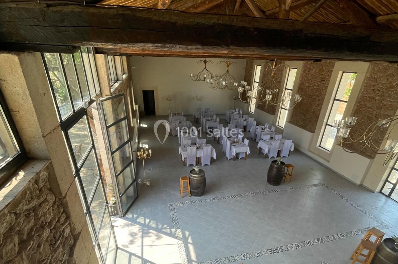 Salle de réception lumineuse avec tables dressées, grandes fenêtres, murs en pierre et poutres apparentes.