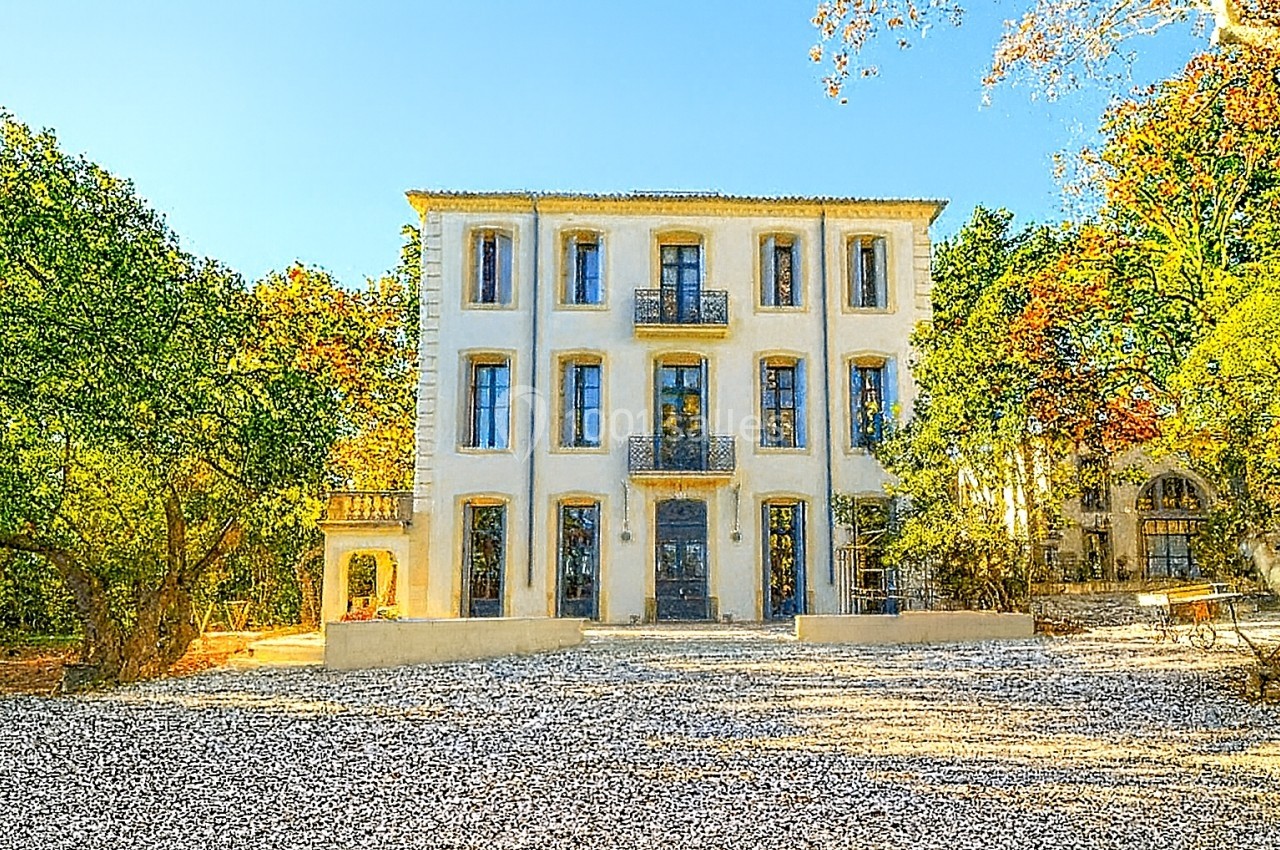 Façade d'un bâtiment ancien à trois étages entouré d'arbres, avec une cour en gravier au premier plan.