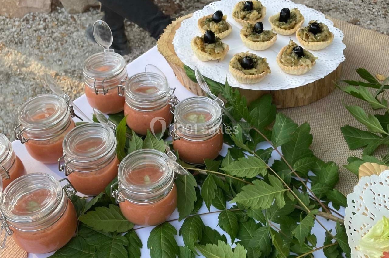 Petits pots de soupe froide et tartelettes aux olives noires disposés sur une table décorée de feuilles vertes.