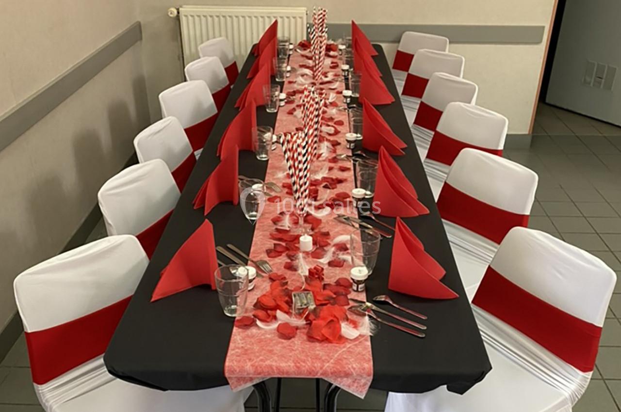 Table décorée avec une nappe noire, des serviettes rouges, des pétales et des bougies, entourée de chaises blanches ornées…