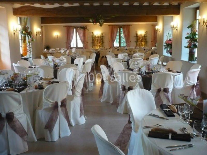 Salle de réception décorée avec des tables rondes dressées, nappes blanches et nœuds en tissu sur les chaises.