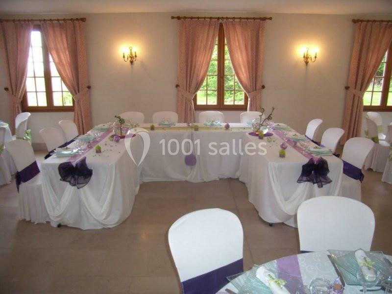 Salle décorée pour un événement avec tables en U, nappes blanches, rubans violets et fenêtres encadrées de rideaux roses.