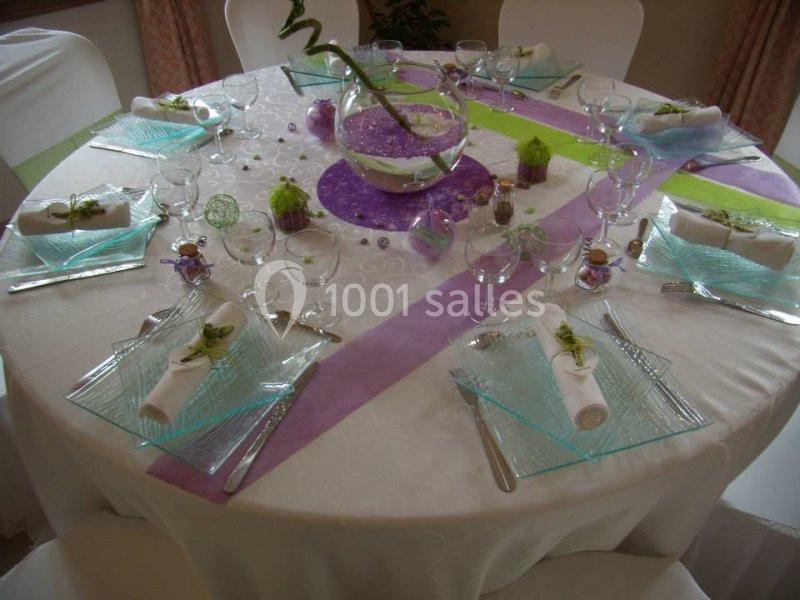 Table ronde décorée avec une nappe blanche, des serviettes pliées, des assiettes en verre et une décoration centrale colorée.