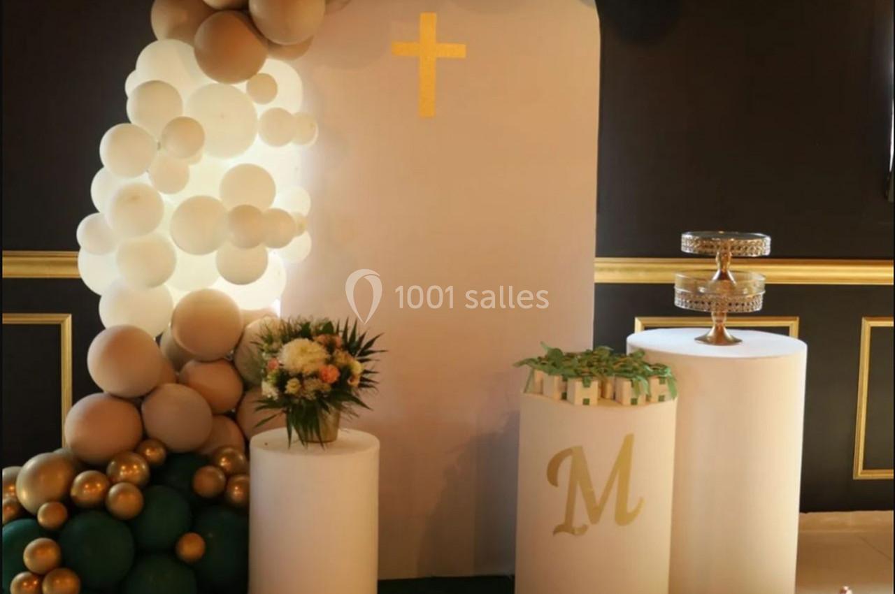 Décoration avec ballons, croix dorée, colonnes blanches, bouquet de fleurs et présentoir à gâteaux sur fond sombre.