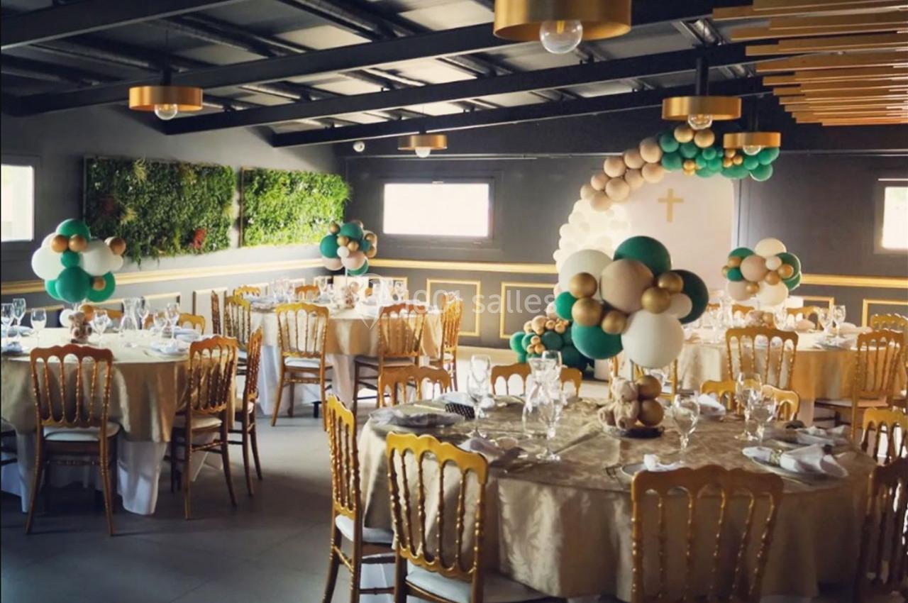 Salle de réception décorée avec des ballons verts, blancs et dorés, tables dressées et mur végétal en arrière-plan.
