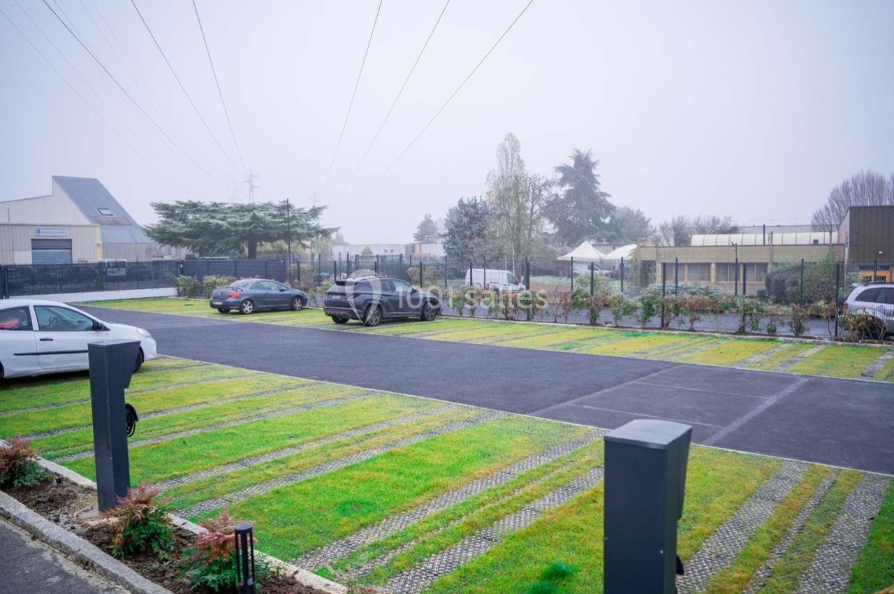 Parking extérieur avec des places alternant pavés et gazon, entouré de bâtiments et d'arbres par temps brumeux.