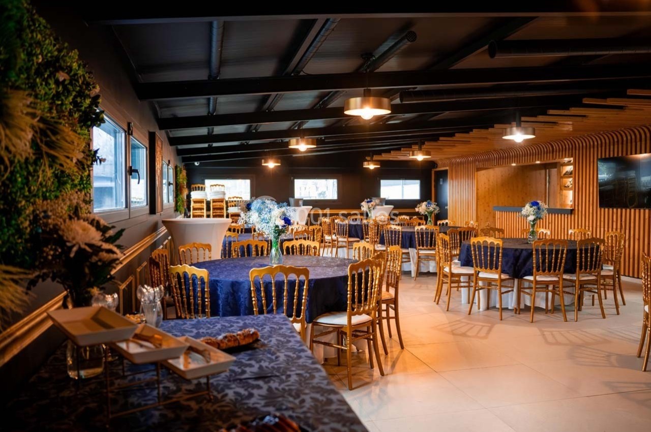 Salle de réception élégante avec tables rondes dressées de nappes bleues, chaises dorées et décorations florales.