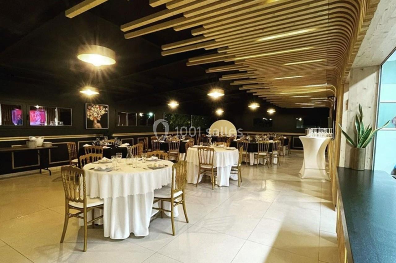 Salle de réception élégante avec tables rondes dressées, chaises dorées et éclairage chaleureux.