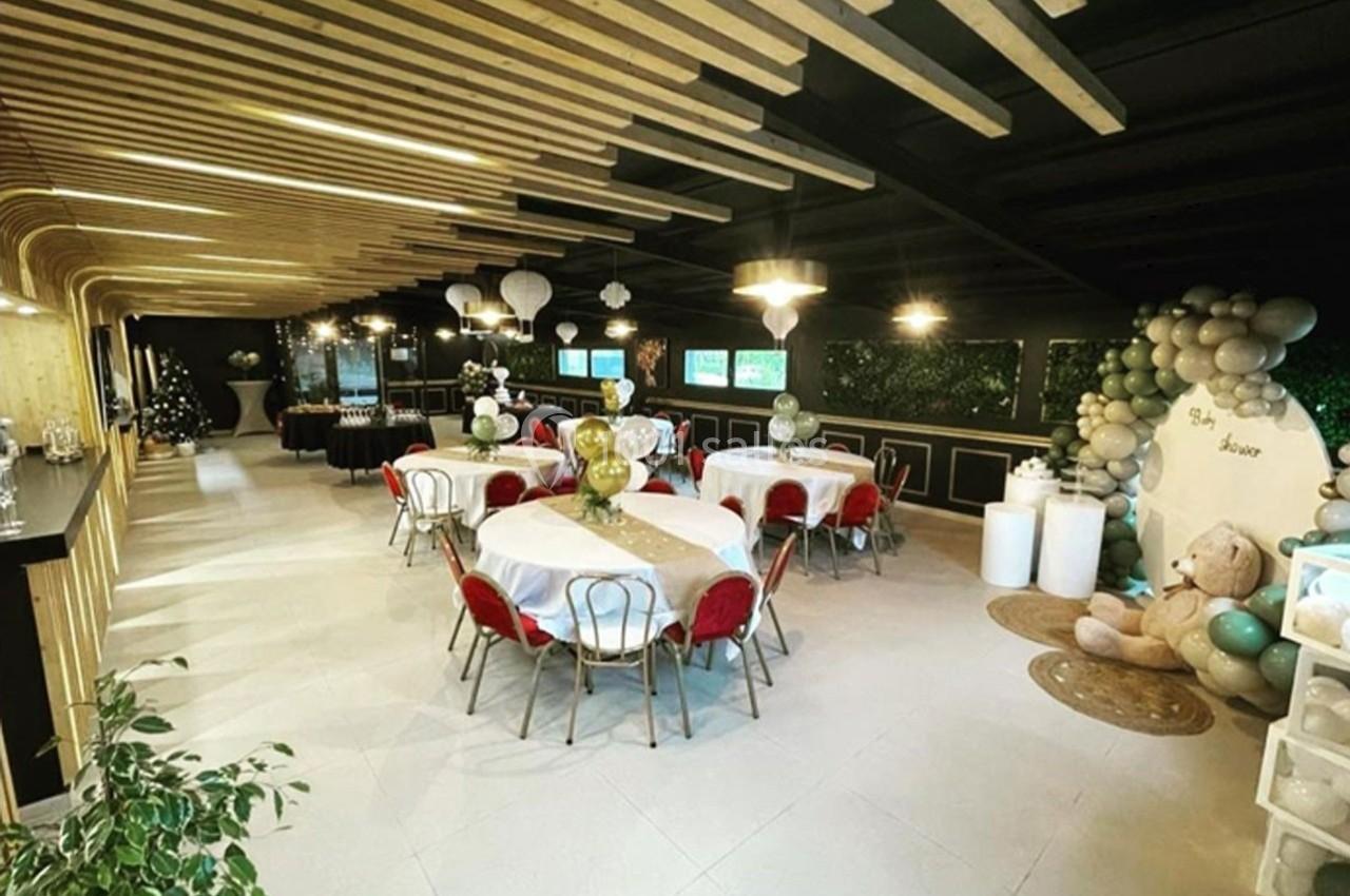 Salle décorée pour un événement avec tables rondes, chaises rouges, ballons, et un espace dédié à un ours en peluche.