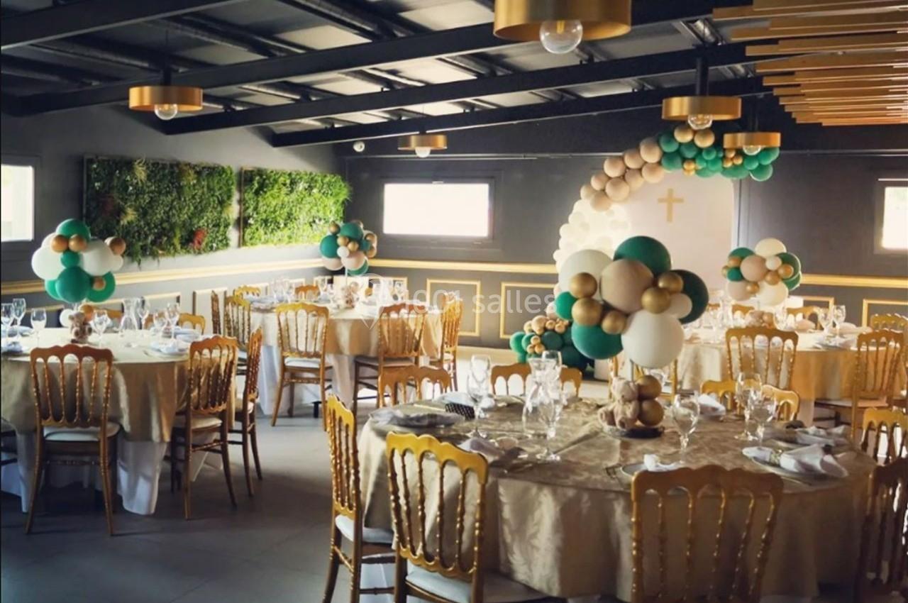 Salle de réception décorée avec des ballons verts et dorés, tables dressées avec nappes beiges et chaises dorées.