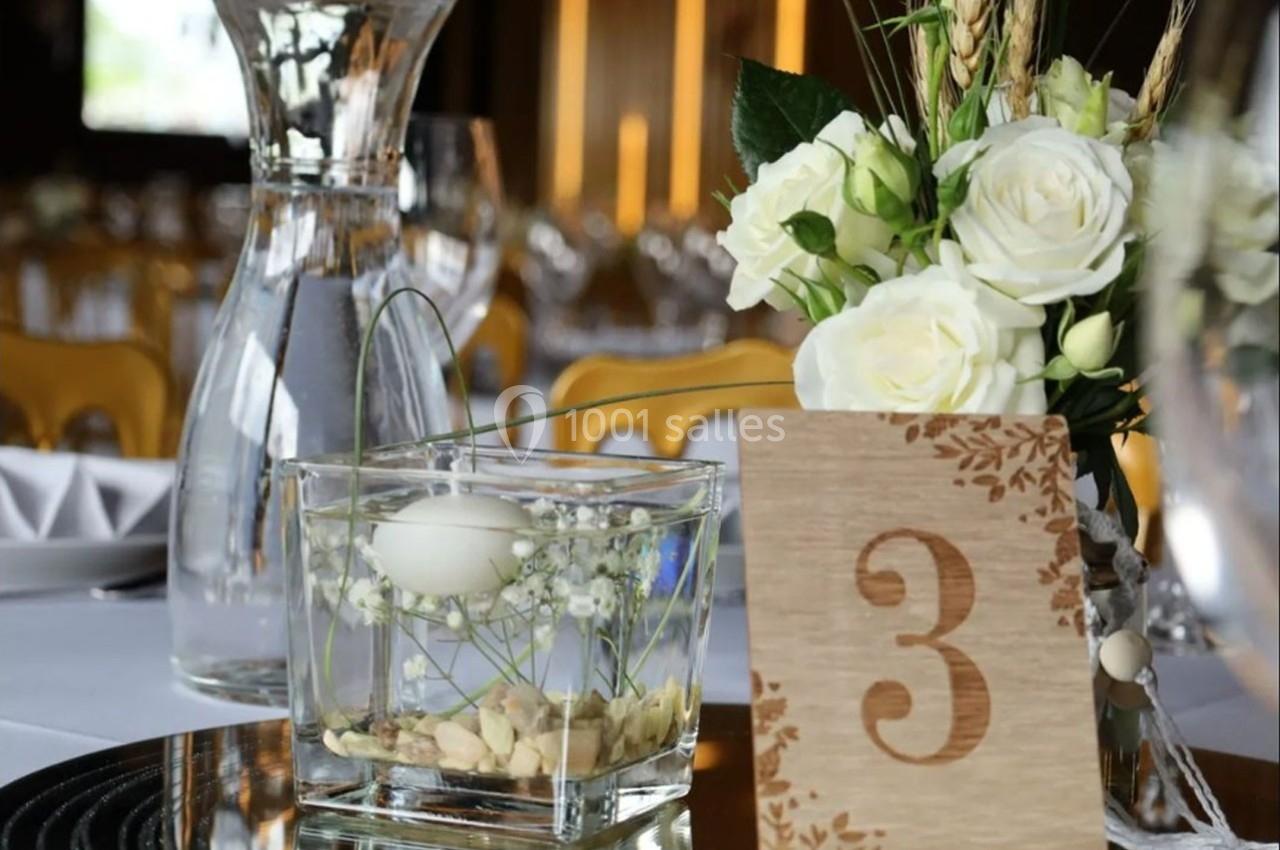 Centre de table avec une carafe d'eau, un vase contenant une bougie flottante, des fleurs blanches et un numéro 3 en bois.