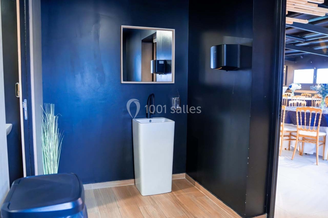 Lavabo moderne avec robinet noir sur fond de mur sombre, situé près d'une poubelle et d'un distributeur mural.