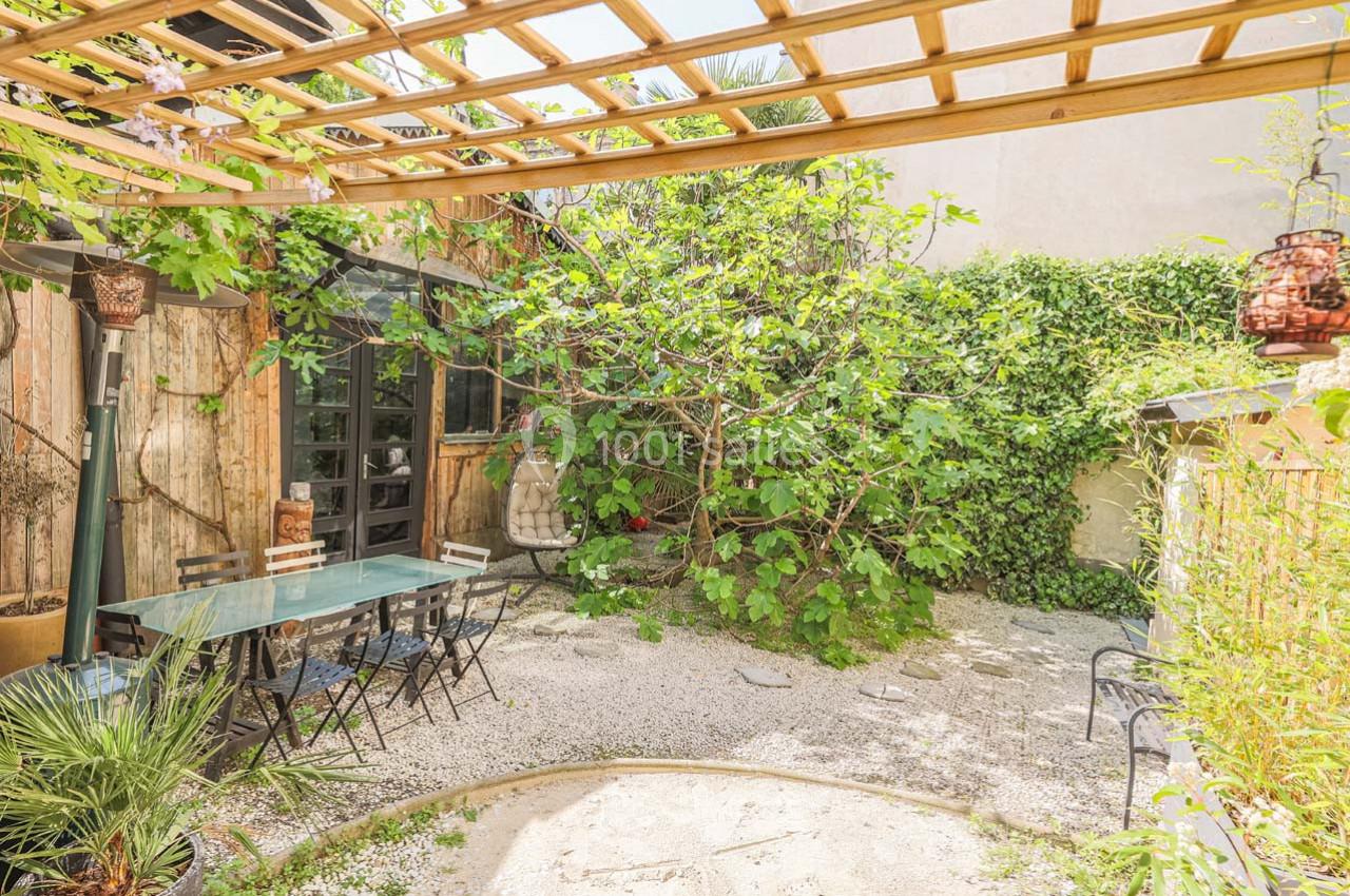 Terrasse extérieure avec table en verre, chaises noires, pergola en bois, végétation dense et sol en gravier.