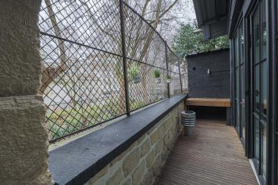 Balcon étroit avec sol en bois, mur en pierre, grillage métallique et vue sur un jardin arboré.