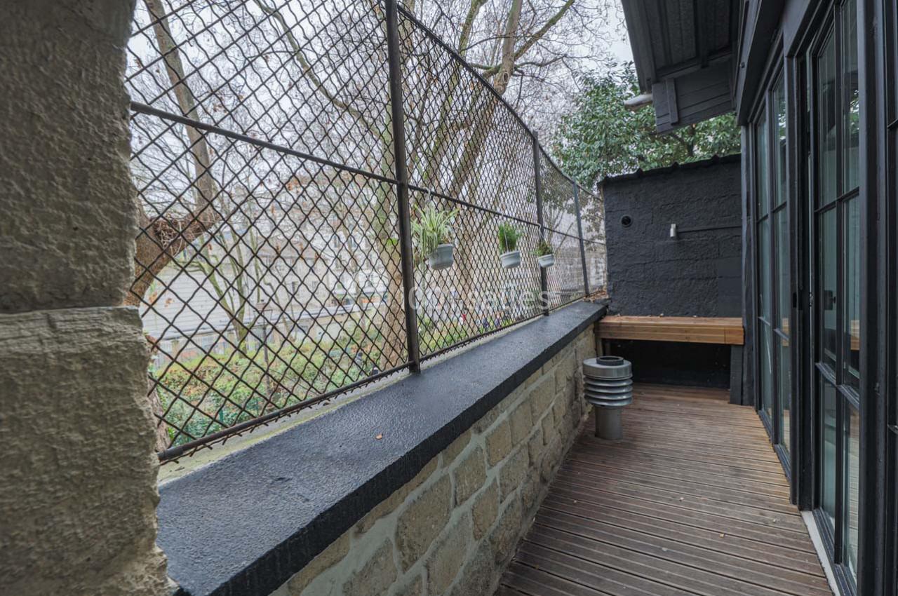 Balcon étroit avec sol en bois, mur en pierre, grillage métallique et vue sur un jardin arboré.