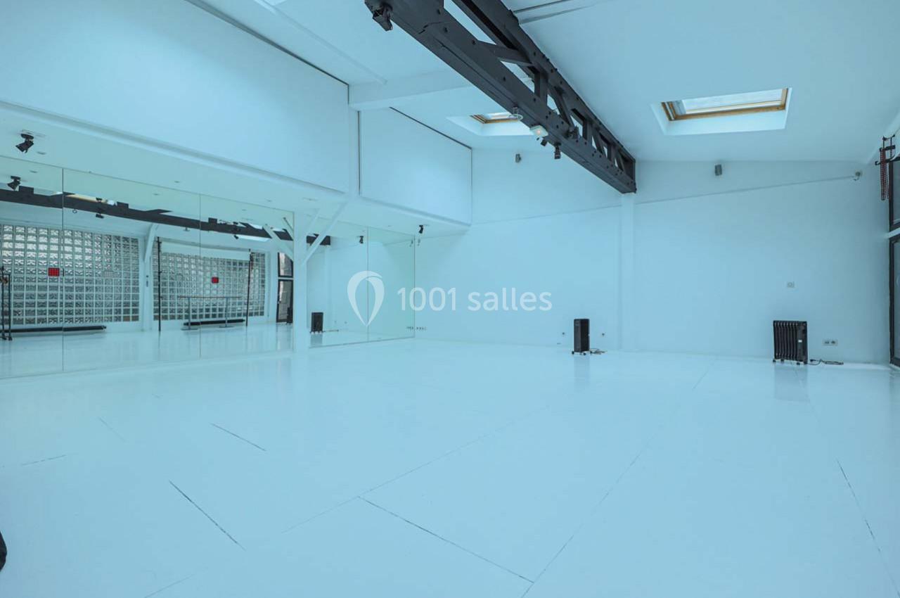 Salle lumineuse avec murs blancs, sol clair, miroirs sur un côté et structure métallique au plafond.