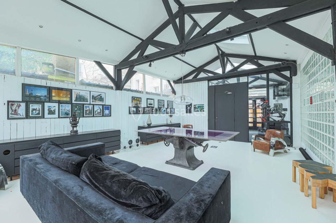 Salle lumineuse avec poutres apparentes, canapé noir, table de ping-pong et murs décorés de cadres.