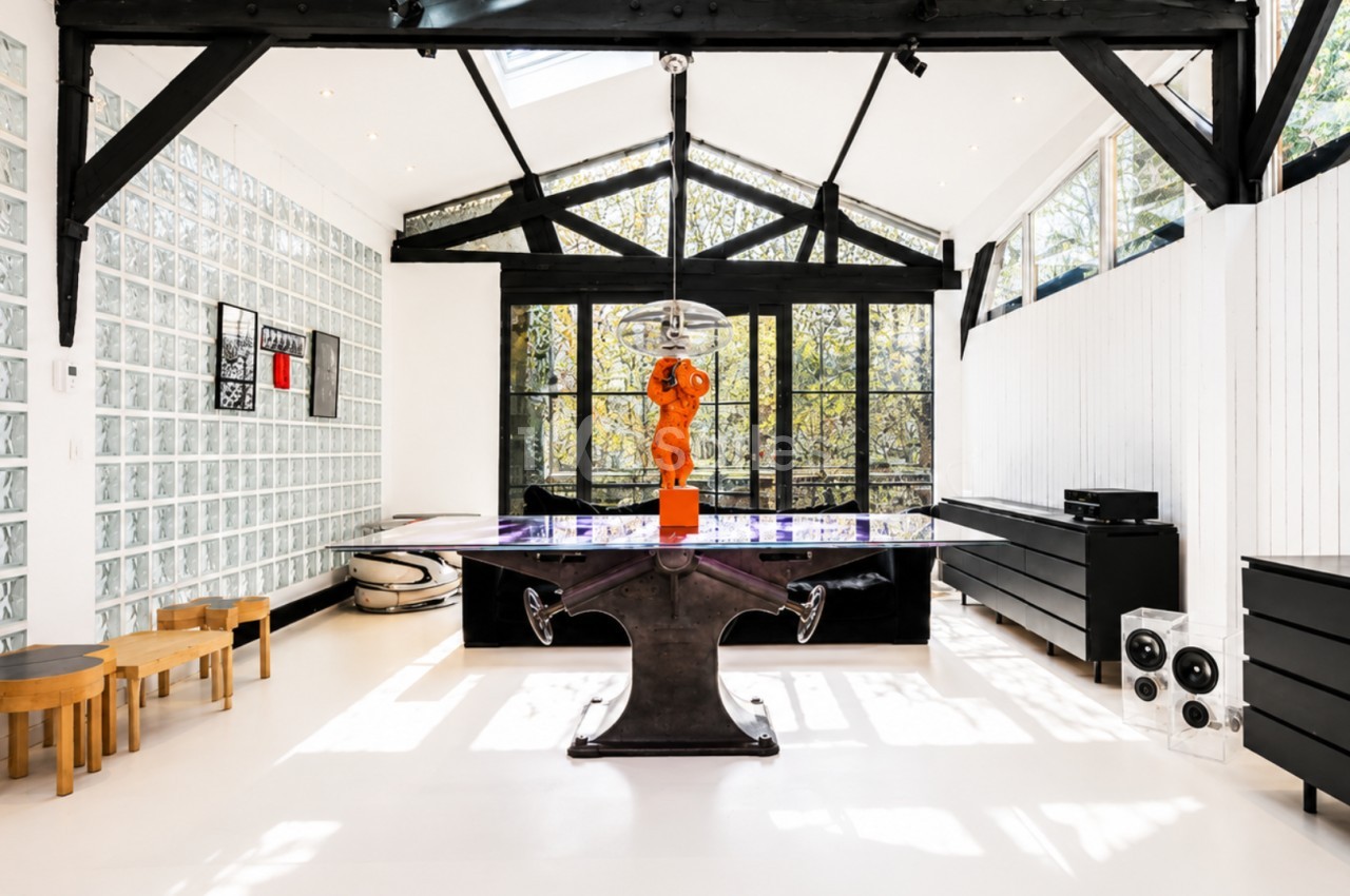 Salle lumineuse avec verrière, table design noire, sculpture orange centrale, murs blancs et mobilier moderne.