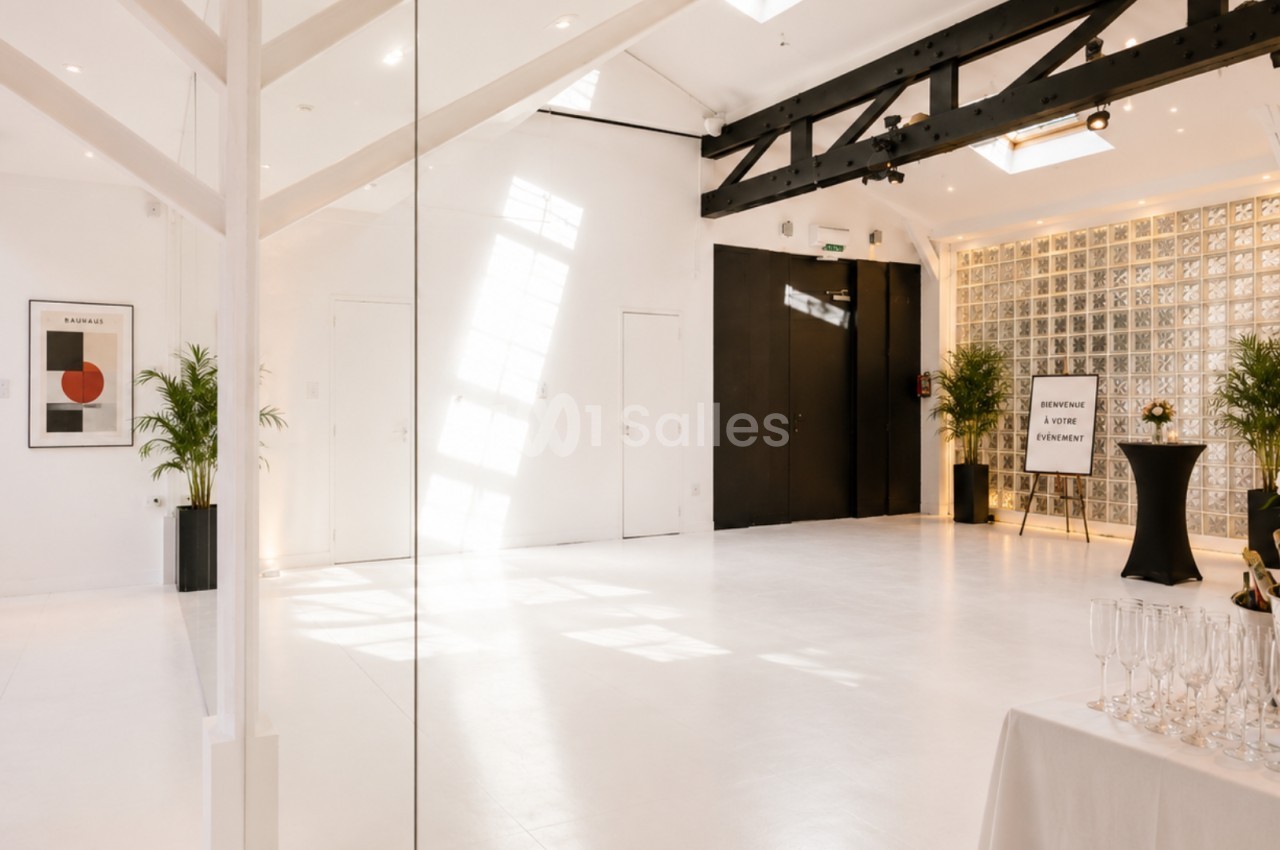 Salle lumineuse avec sol blanc, poutres noires apparentes, plantes décoratives et table dressée pour un événement.