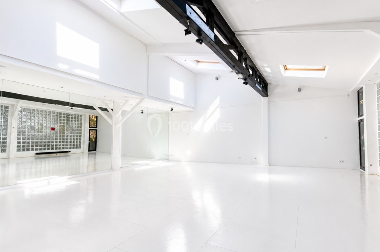 Salle lumineuse avec murs blancs, sol clair, miroirs, verrière et poutres noires apparentes.