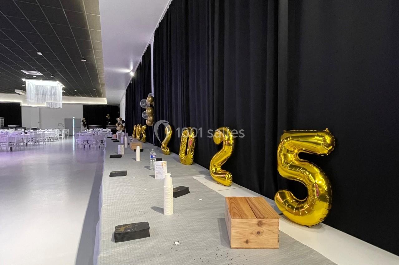 Ballons dorés formant ’2025’ alignés sur une table décorée, dans une salle avec rideaux noirs et chaises en arrière-plan.