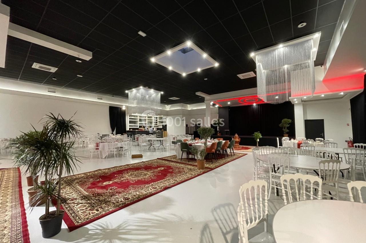 Salle de réception spacieuse avec tables rondes, chaises blanches, tapis rouges et scène éclairée en arrière-plan.