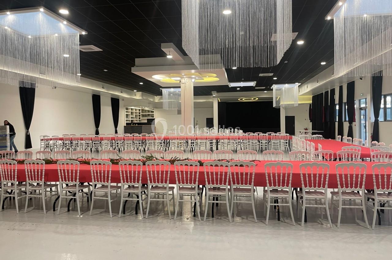 Salle de réception avec tables rectangulaires alignées, nappes rouges, chaises blanches et décorations suspendues.