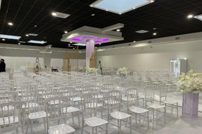 Salle de réception décorée avec des chaises blanches alignées et des arrangements floraux, prête pour un événement.