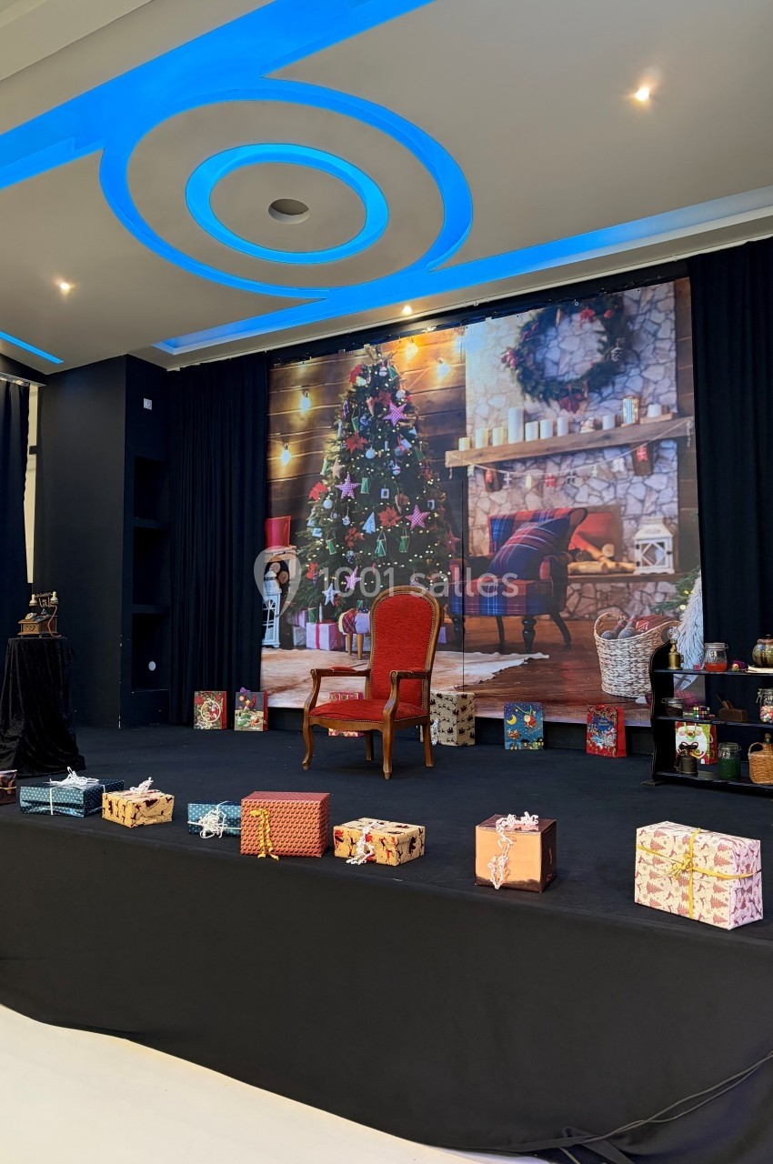 Scène décorée avec un sapin de Noël illuminé, une cheminée, un fauteuil rouge et des cadeaux emballés au premier plan.