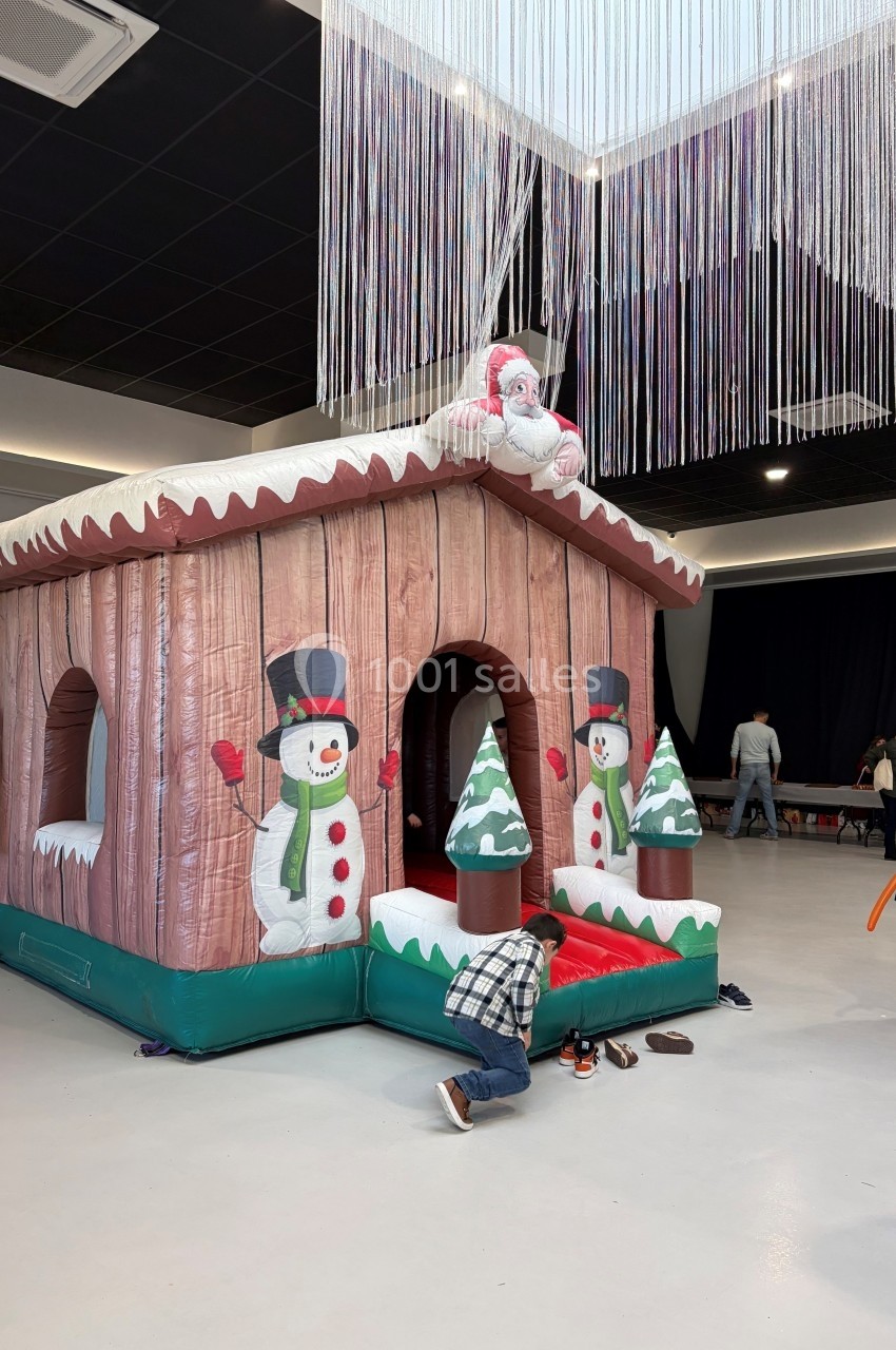 Structure gonflable en forme de chalet décoré de bonhommes de neige et de sapins, installée dans un espace intérieur.