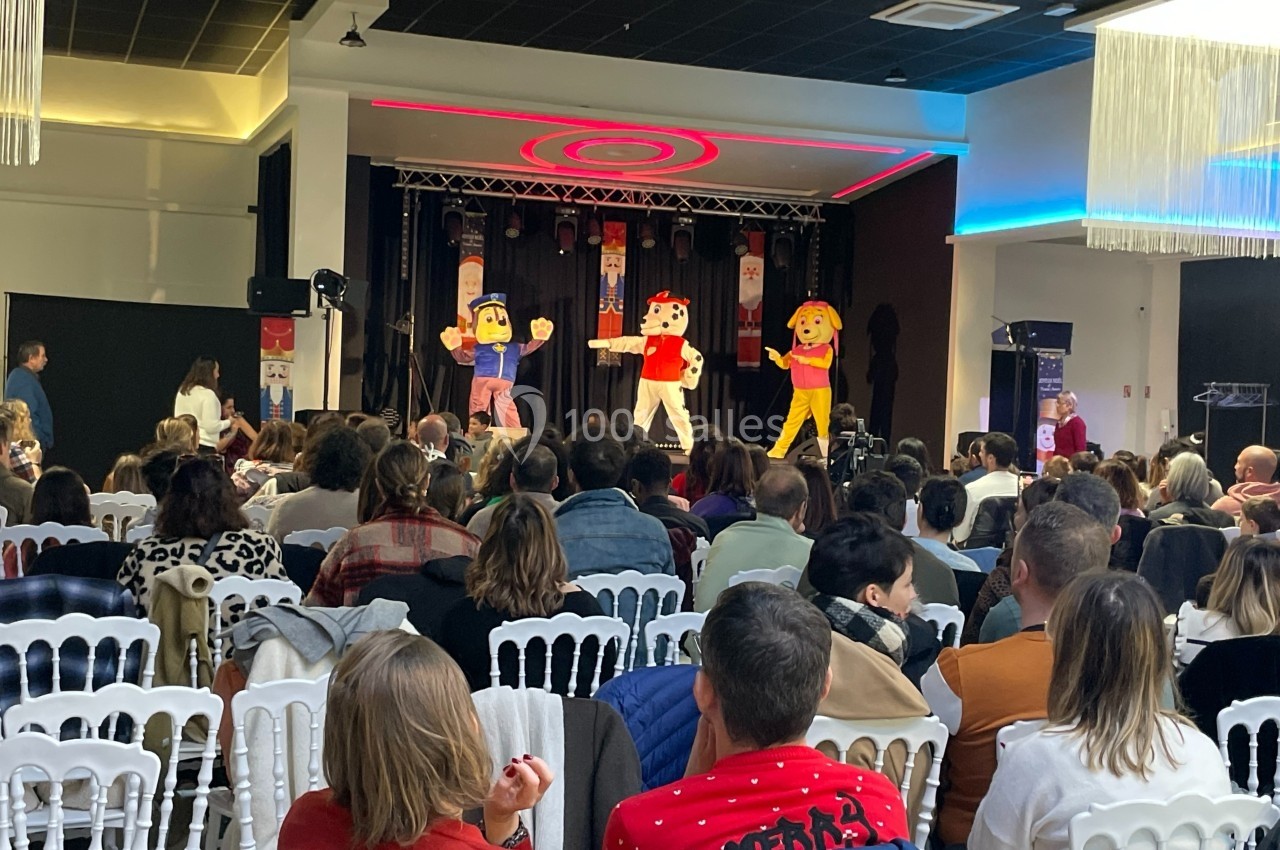Des mascottes colorées dansent sur scène devant un public assis dans une salle de spectacle.