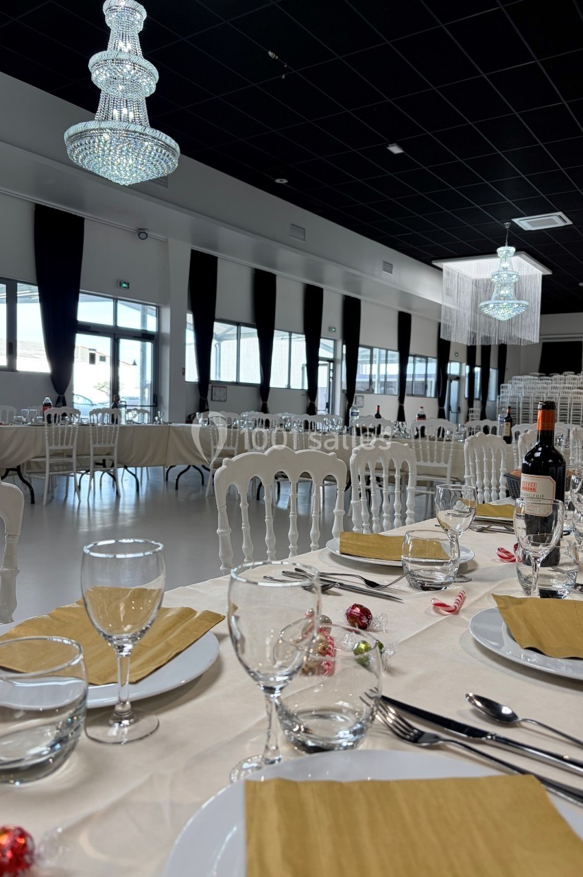 Salle de réception élégante avec tables dressées, nappes blanches, serviettes dorées, verres et chandeliers suspendus.