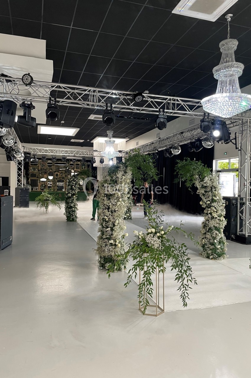 Salle décorée pour un événement avec des arches fleuries, des lustres suspendus et une structure métallique éclairée.