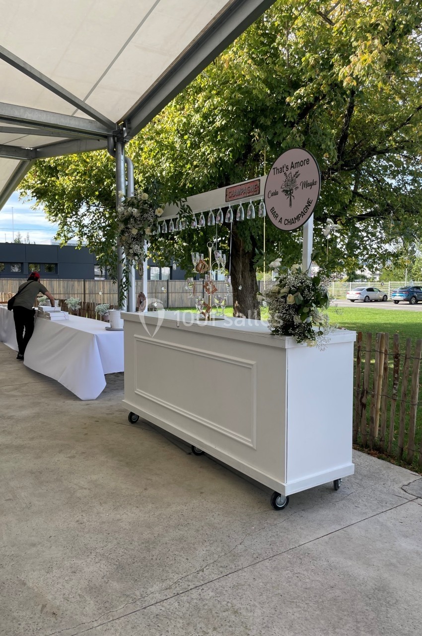 Bar mobile blanc décoré de fleurs et de verres suspendus, installé en extérieur sous une tente près d'un espace arboré.