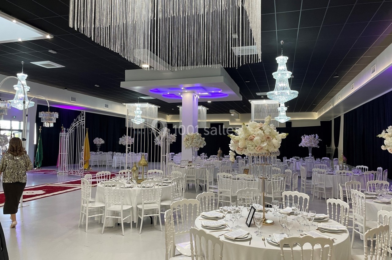 Salle de réception élégante avec tables dressées, chaises blanches, décor floral et lustres suspendus.