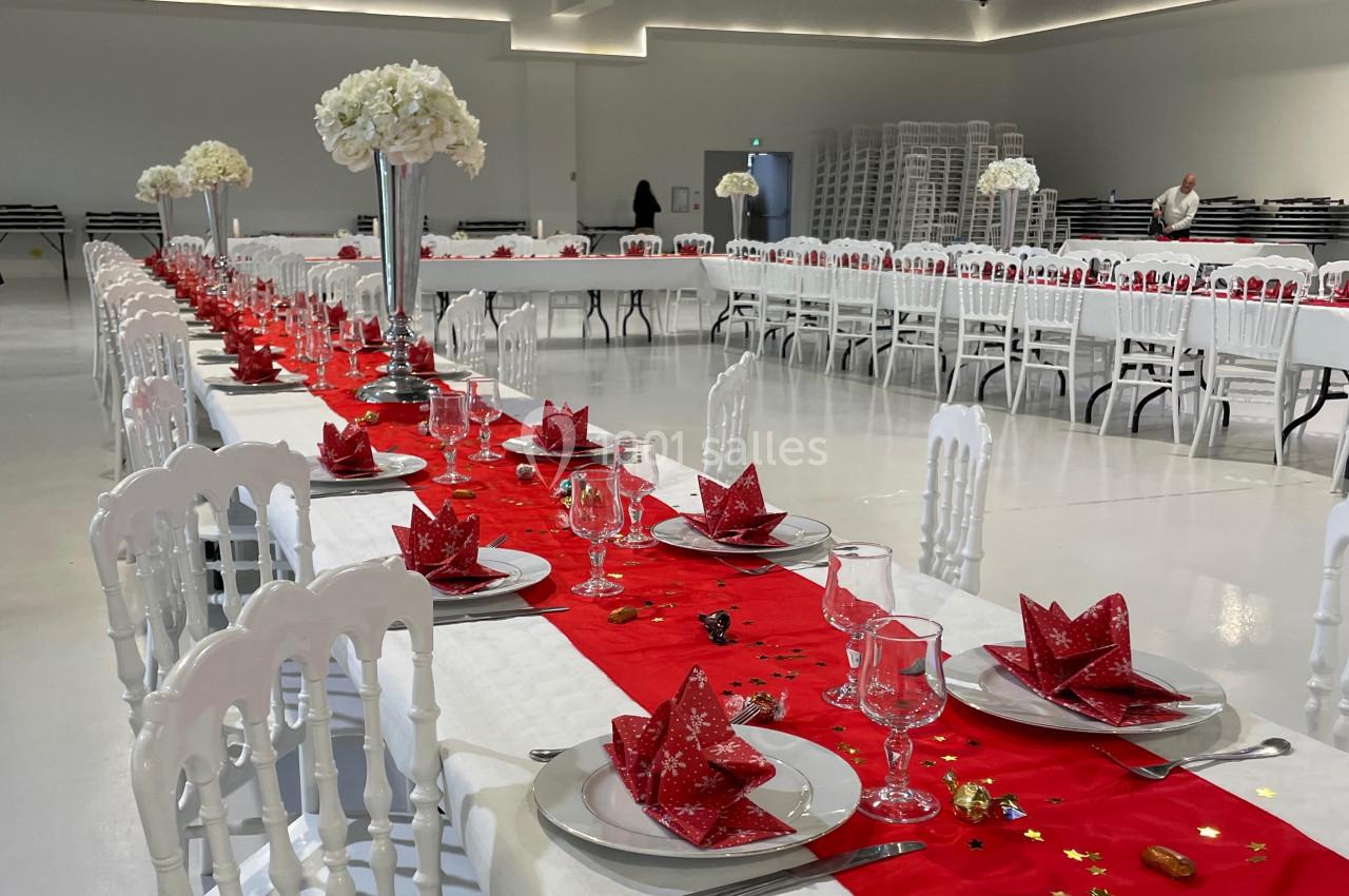 Grande salle décorée pour un événement, avec longues tables dressées, nappes rouges et centres de table floraux.