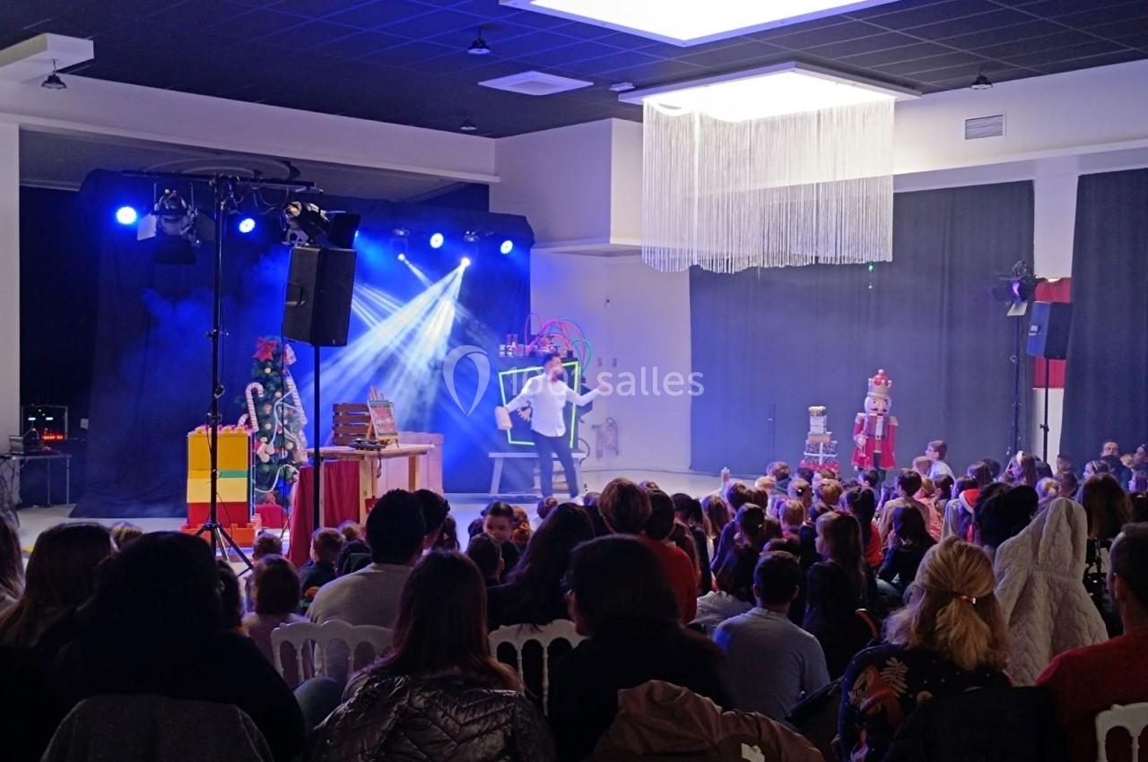 Un magicien se produit sur scène devant un public assis, éclairé par des projecteurs dans une salle sombre.