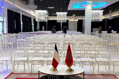Salle de réception avec des rangées de chaises blanches, tapis rouge au premier plan et drapeaux français et marocain sur…