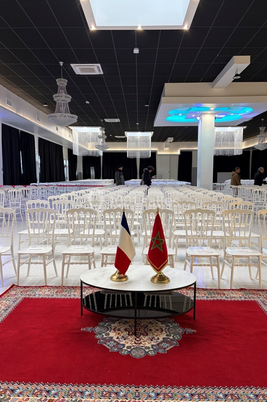 Salle de réception avec des rangées de chaises blanches, tapis rouge au premier plan et drapeaux français et marocain sur…