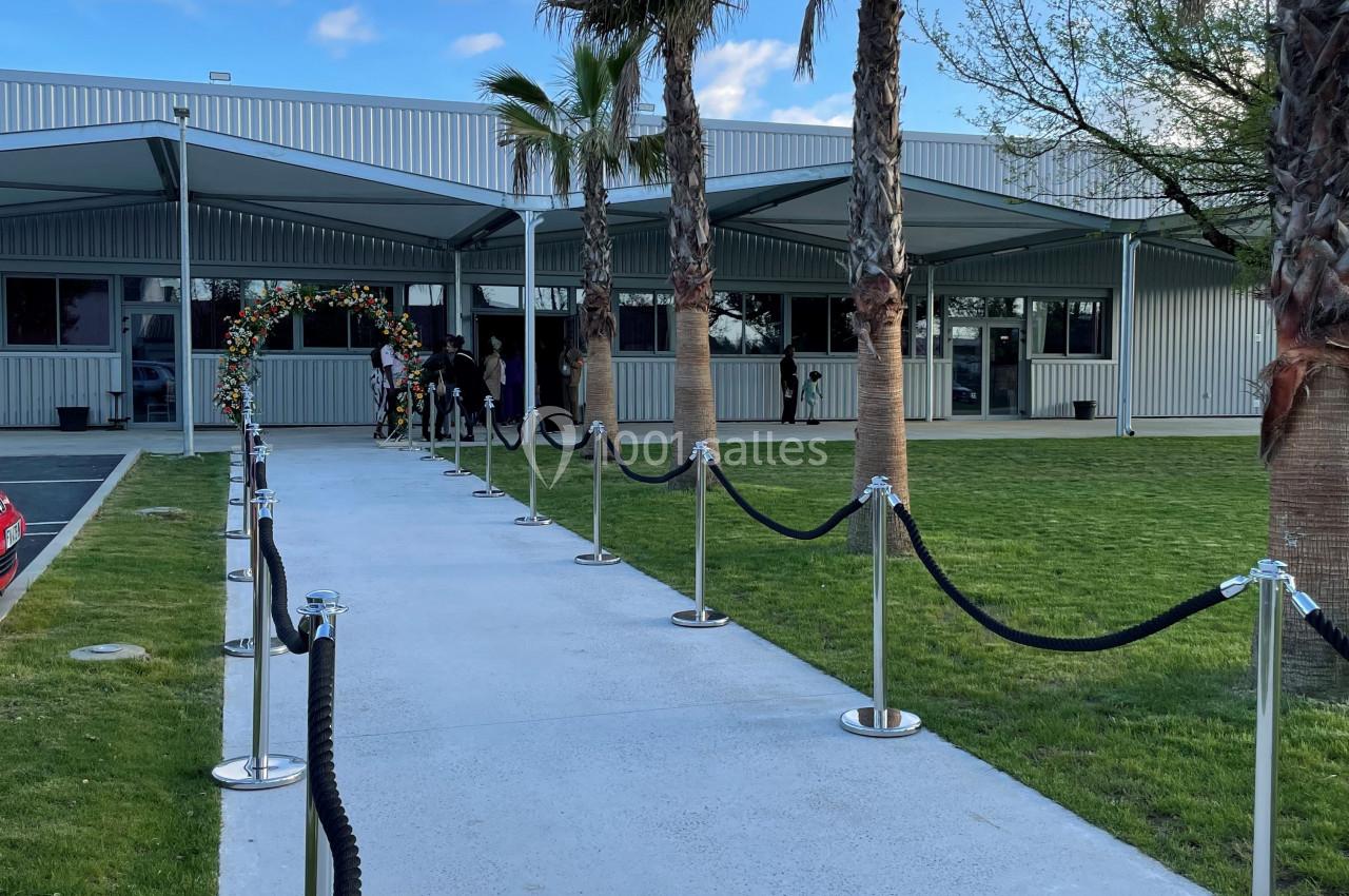 Allée bordée de cordons menant à un bâtiment moderne avec palmiers et ciel partiellement nuageux.