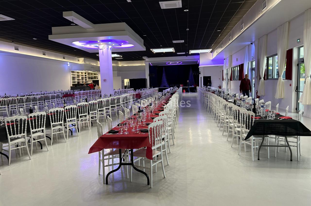Grande salle de réception avec tables alignées, nappes rouges et noires, chaises blanches et éclairage moderne.