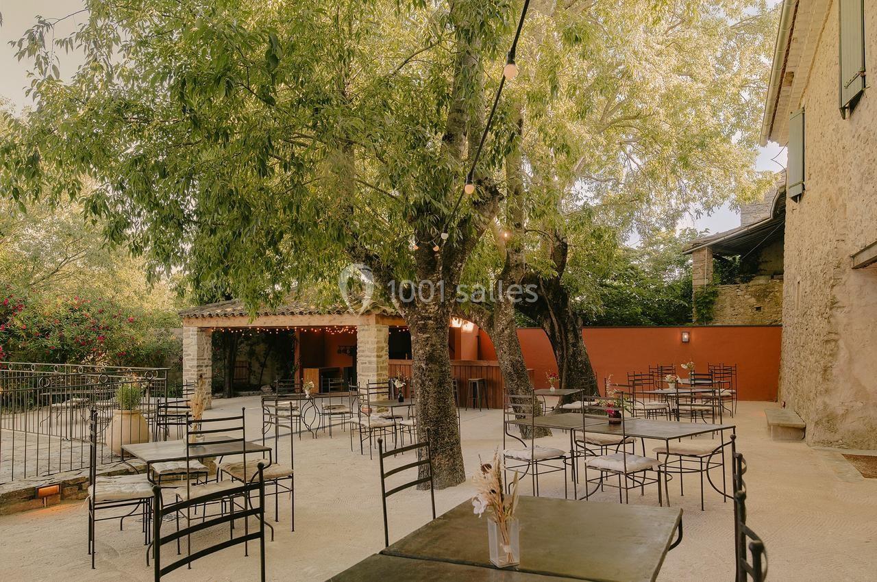 Terrasse extérieure avec tables et chaises sous des arbres, près d'un bâtiment en pierre et d'un bar couvert.