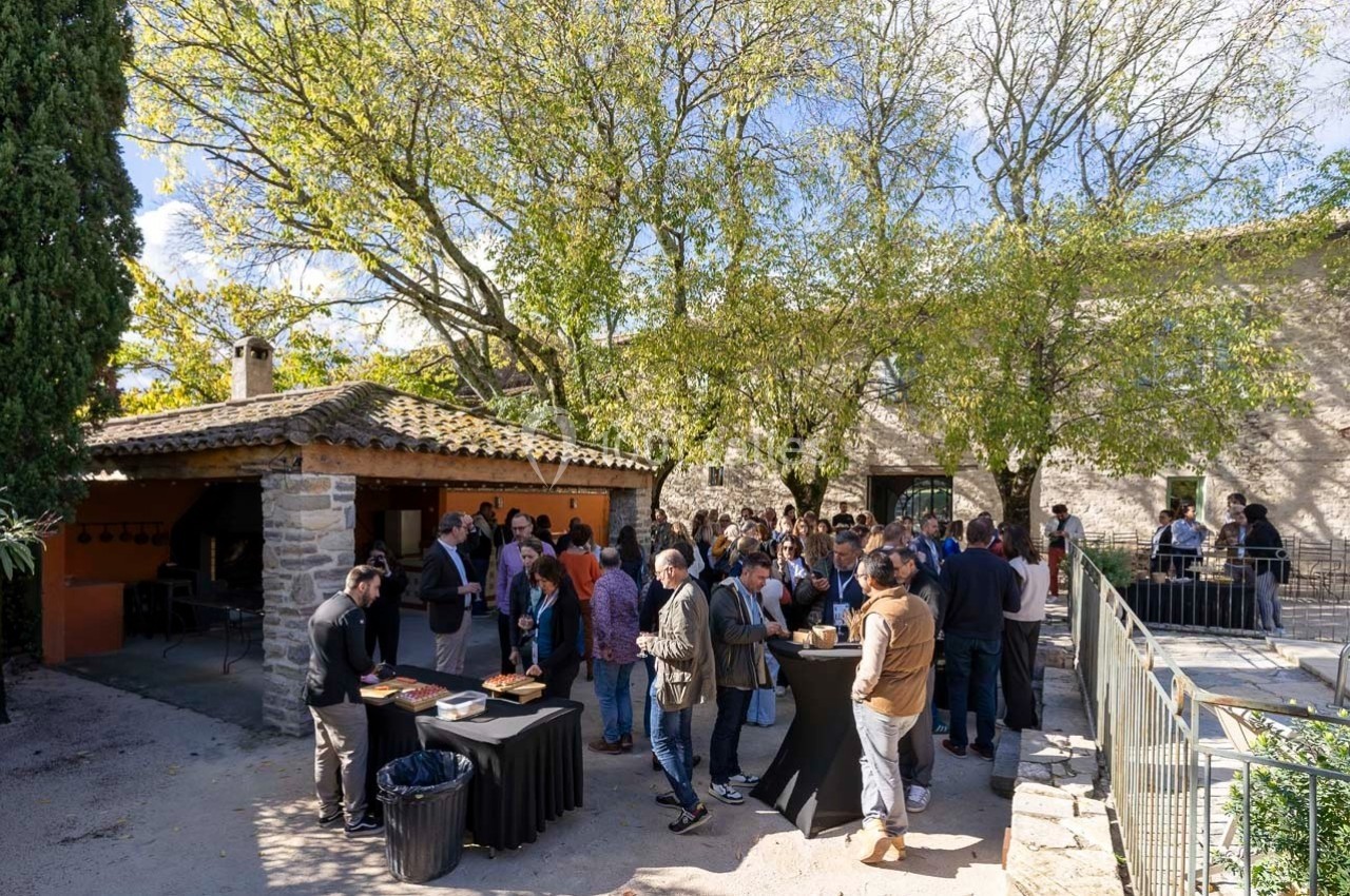 Groupe de personnes rassemblées en extérieur près d'un buffet, dans une cour entourée d'arbres et de bâtiments en pierre.