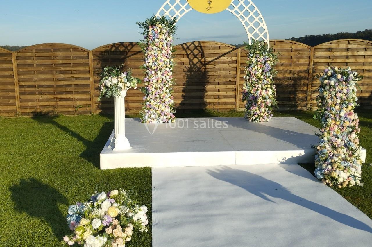 Arche de mariage décorée de fleurs pastel sur une estrade blanche, entourée de pelouse et de ciel dégagé.